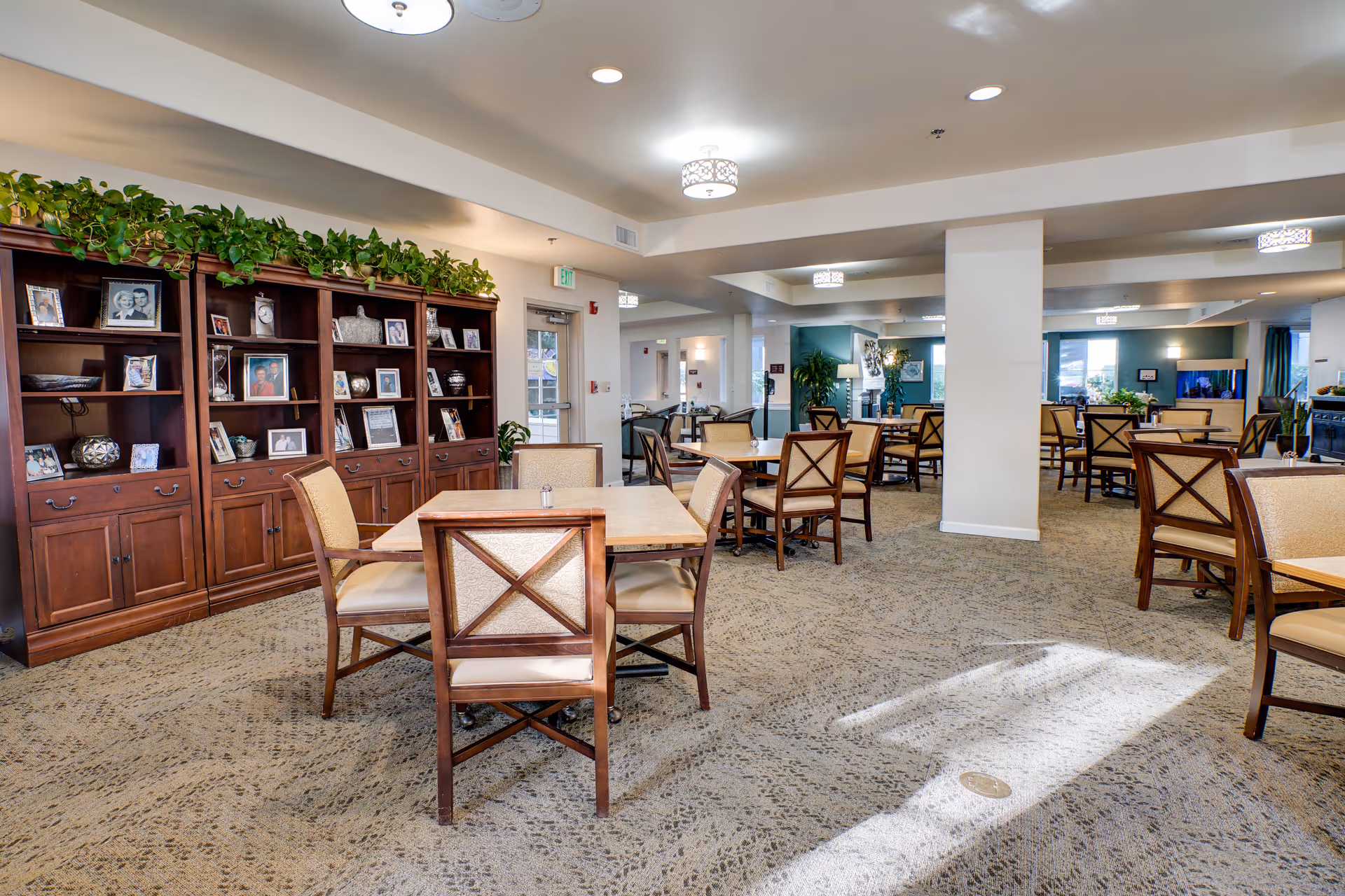 Spacious retirement community dining room with multiple tables and chairs, a large wooden bookshelf displaying framed photos, and soft overhead lighting.
