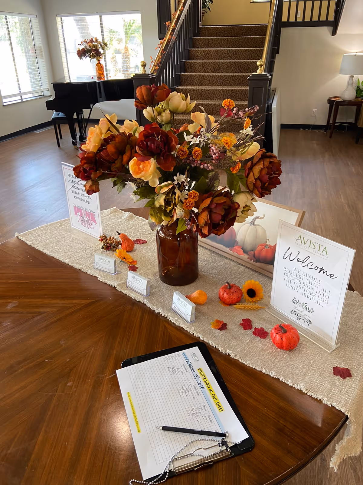 A wooden table with a beige runner decorated with artificial autumn flowers in a brown vase, small pumpkins, and fall leaves. On the table is a visitor sign-in sheet on a clipboard with a pen. Behind the table is a staircase with carpeted steps and a black railing, a grand piano near windows letting in natural light, and a small side table with a lamp and plant. Signs on the table include a welcome message and a breast cancer awareness notice.