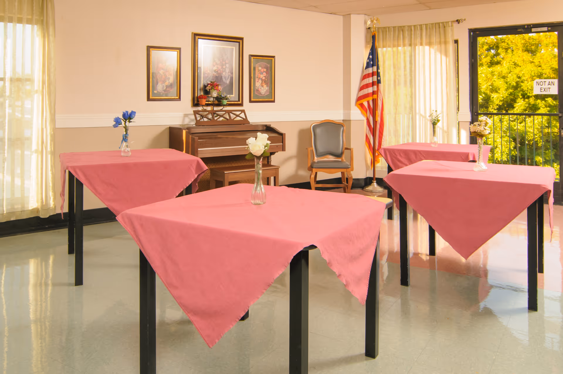 A communal dining/activity room with square tables draped in pink tablecloths, small vases of flowers, a piano and an American flag by the windows.