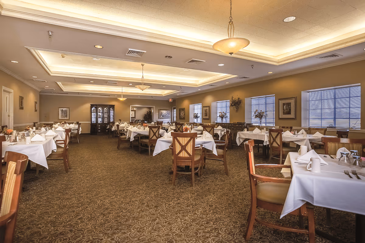 A spacious dining room with multiple tables covered in white tablecloths, each set with napkins, cups, and silverware. The room features warm lighting from ceiling fixtures, large windows with blinds, framed artwork on the walls, and a carpeted floor. Wooden chairs with cushioned seats are arranged around the tables.