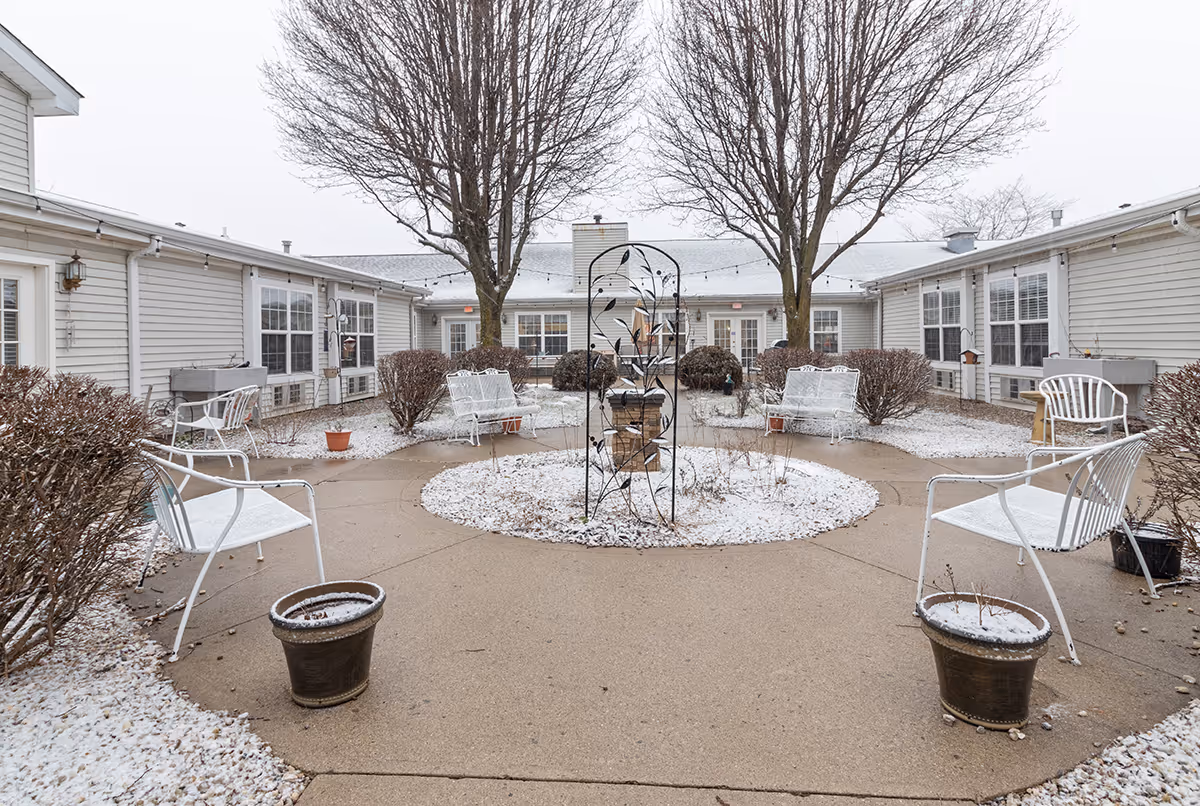 Outdoor courtyard area at Campbell Place with a circular concrete pathway surrounding a central garden bed with a metal trellis. There are leafless trees and several white metal benches and chairs placed around the courtyard. The ground and plants have a light dusting of snow, and the surrounding building has beige siding with multiple windows and doors.