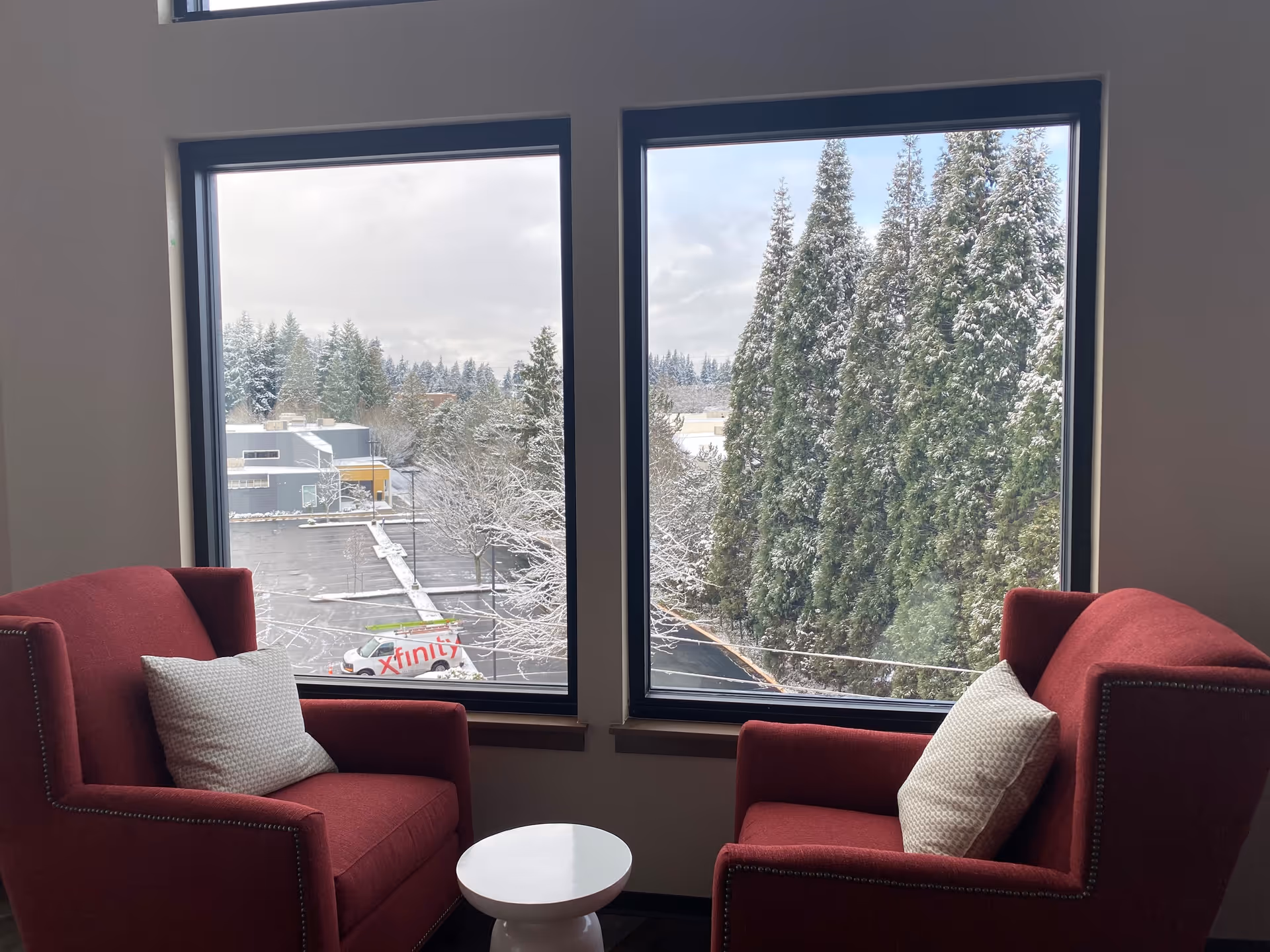 Two red armchairs with white patterned pillows facing each other with a small white round table between them, positioned in front of two large windows showing a snowy outdoor scene with tall evergreen trees and a parking lot.