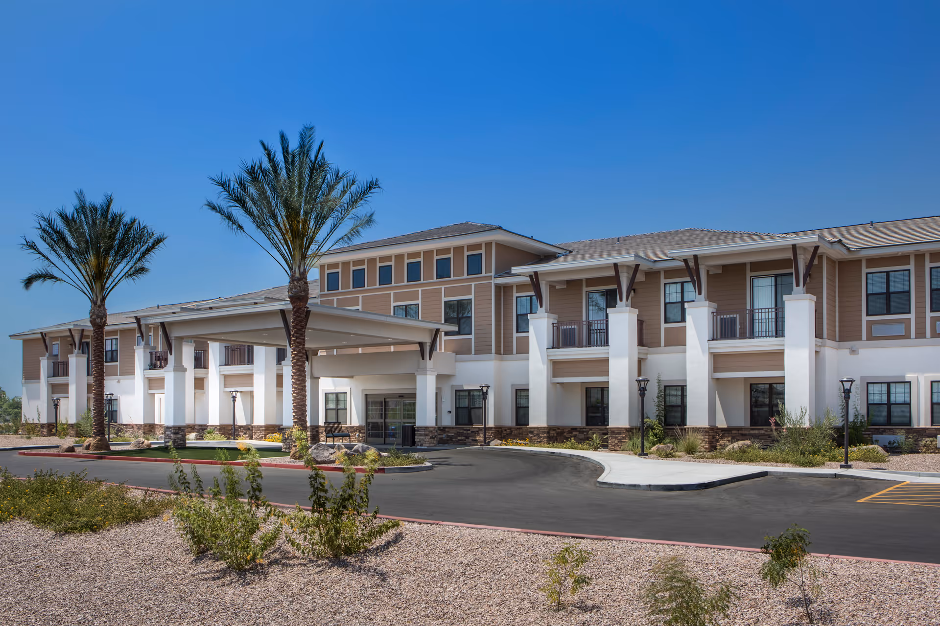Front exterior of a two-story beige and white building with a covered entrance, palm trees, and landscaped gravel beds.
