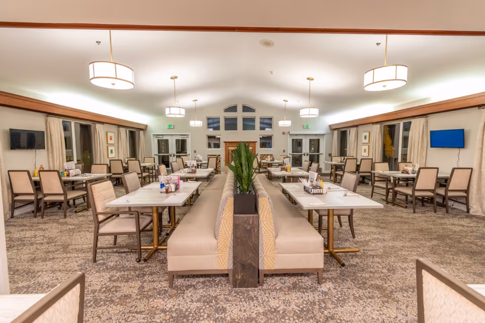 A spacious dining room with multiple tables and chairs arranged neatly. The room features a long cushioned bench with a planter in the center, pendant lights hanging from the ceiling, large windows with curtains, and two wall-mounted televisions.