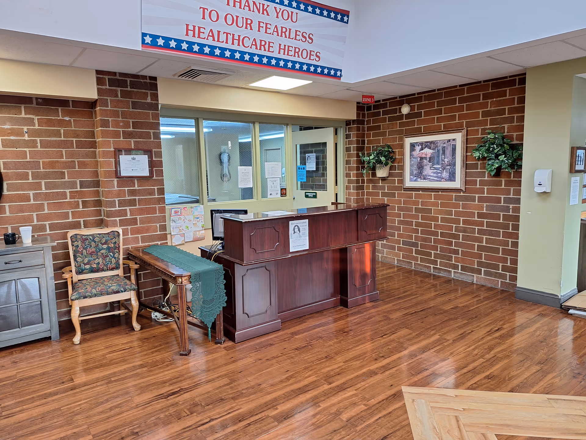 Reception area with a wooden desk, a floral upholstered chair, a small table with a green runner, and a brick wall with framed artwork and potted plants. A banner above reads 'Thank you to our fearless healthcare heroes.'