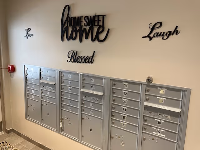 A wall with multiple silver mailboxes numbered from 1 to 30, mounted below decorative black wall words that say 'Love', 'Home Sweet Home', 'Blessed', and 'Laugh'. There is a fire alarm on the left side of the wall and a surveillance camera above the mailboxes.