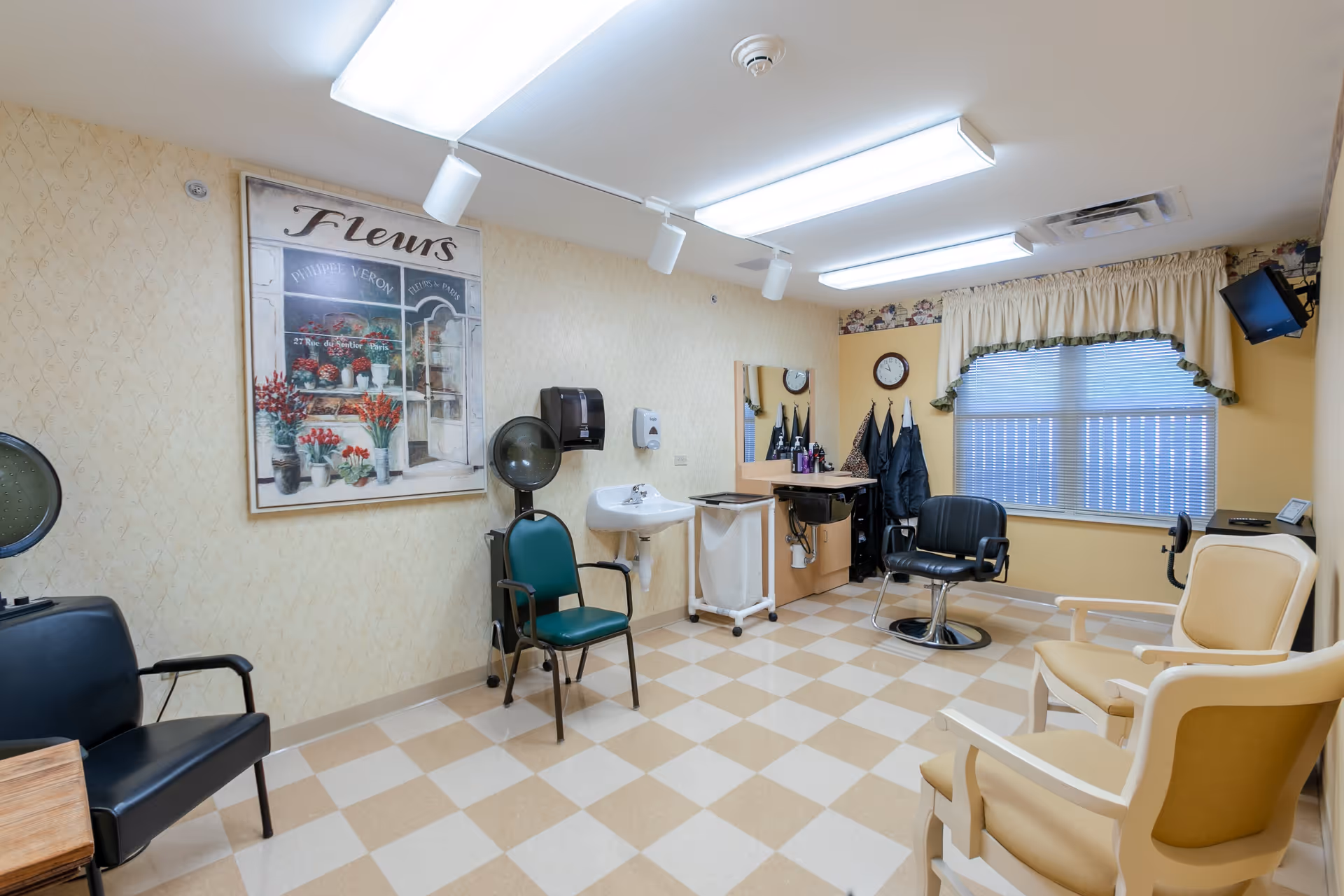 A small salon room with beige and cream checkered flooring, several chairs including salon chairs and waiting chairs, a wall-mounted hair dryer, a sink, a mirror with hair care products, a clock, a window with blinds and a valance, and a large framed picture of flowers on the wall.