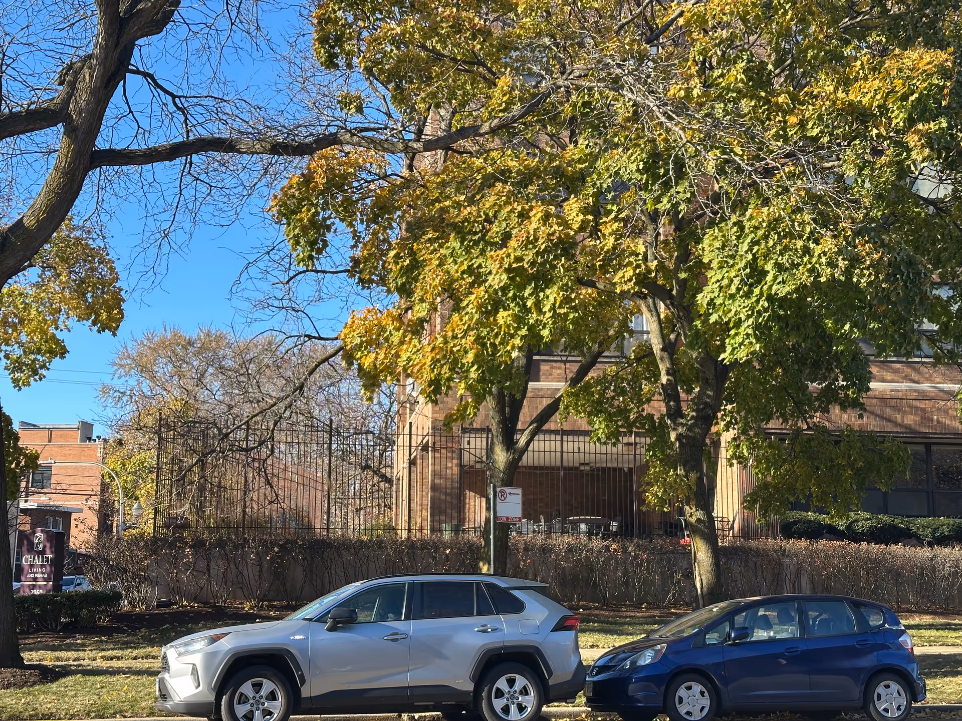 Two parked cars, one silver and one blue, are in front of a brick building partially obscured by trees with green and yellow leaves under a clear blue sky. A sign for Chalet Living And Rehab Center is visible on the left side near the sidewalk.