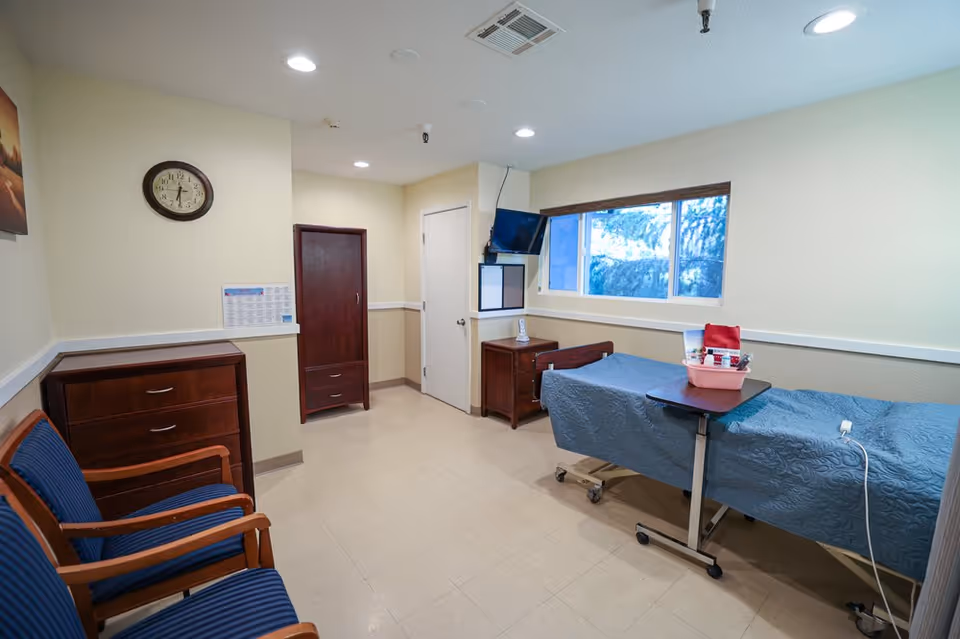 A small, clean patient room with a hospital bed covered in a blue quilt, a rolling overbed table with toiletries, two wooden chairs with blue cushions, a wooden dresser, a wooden wardrobe, a small wooden nightstand with a phone, a wall clock, a window showing trees outside, and a wall-mounted TV.