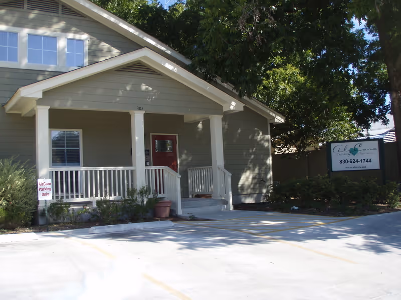 Exterior view of a single-story building with a covered porch supported by white columns. The building is painted light gray with white trim and has a red front door. There is a small garden with bushes in front of the porch and a parking area with a sign that reads 'AlzCare Parking Only'. A larger sign near the right side of the image displays contact information for AlzCare.