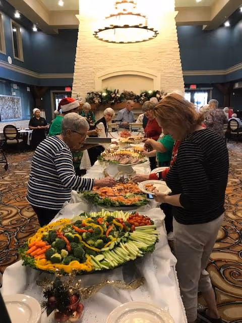 A group of elderly people serving themselves food from a buffet table set up in a large, decorated room with high ceilings and a large stone fireplace. The buffet includes trays of vegetables, fruits, and other dishes. The room has patterned carpet and several tables and chairs in the background.