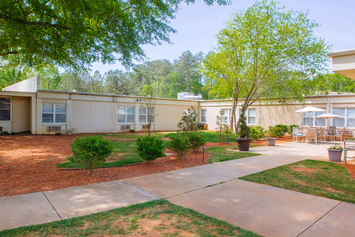 Outdoor courtyard area at Marietta Center for Nursing and Healing with a concrete walkway, green bushes, trees, and patio furniture with umbrellas near the building.