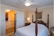 A bedroom with a four-poster bed covered with white bedding, a nightstand with a lamp, a ceiling fan with light, and an open door leading to a bathroom.