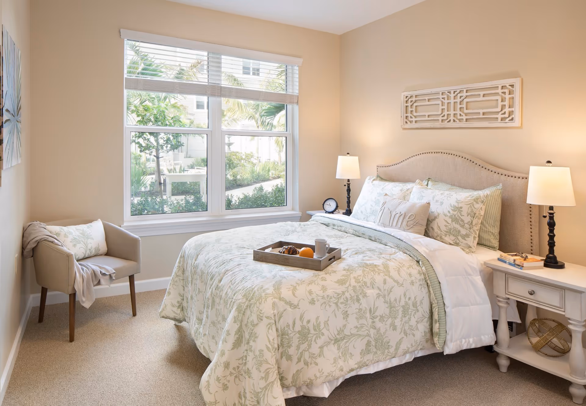 A cozy bedroom with a large window showing greenery outside. The bed is made with floral-patterned bedding in soft green and white tones, with a decorative pillow that says 'Home'. There are two bedside tables with matching lamps, a clock, and some books. A beige armchair with a pillow and a throw blanket is placed near the window. The walls are painted a light beige color, and there is a decorative wall piece above the bed.
