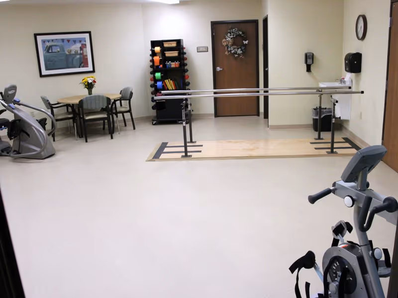 A bright therapy/exercise room with parallel bars, exercise bikes, a small table and chairs, and a rack of fitness equipment.