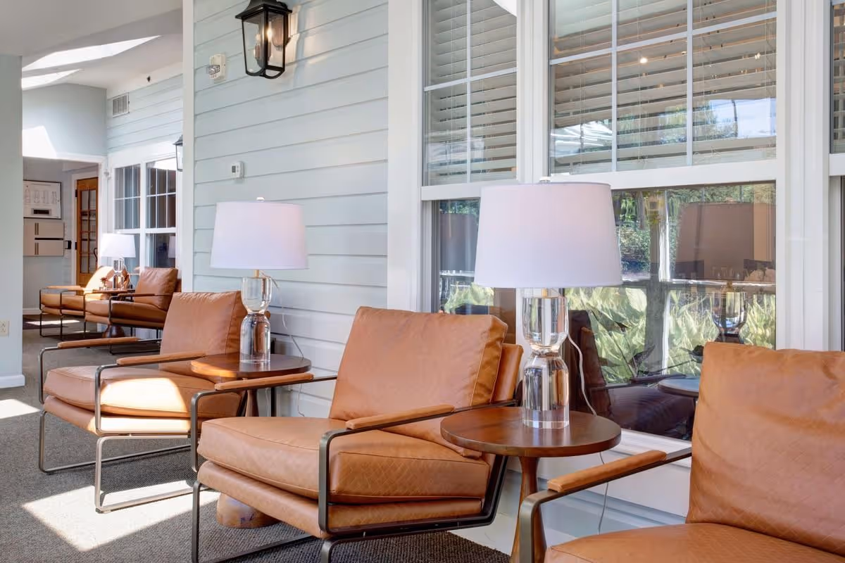 A bright indoor seating area with several tan leather armchairs arranged along a wall with large windows. Each chair is paired with a small round wooden side table holding a modern glass lamp with a white shade. The walls are painted light blue with horizontal paneling, and natural light fills the space.