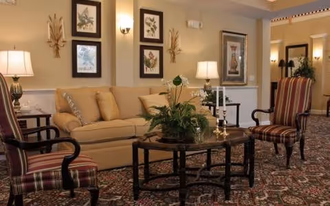 A cozy living room area with a beige sofa, two striped armchairs, a dark wooden coffee table with a green plant and candlesticks, framed botanical artwork on the walls, and warm lighting from table lamps and wall sconces.