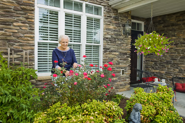An elderly woman tending to a garden with blooming red flowers in front of a stone exterior wall of a building. There is a window with white shutters behind her and a hanging flower basket to the right. A small patio area with chairs and a table is also visible.