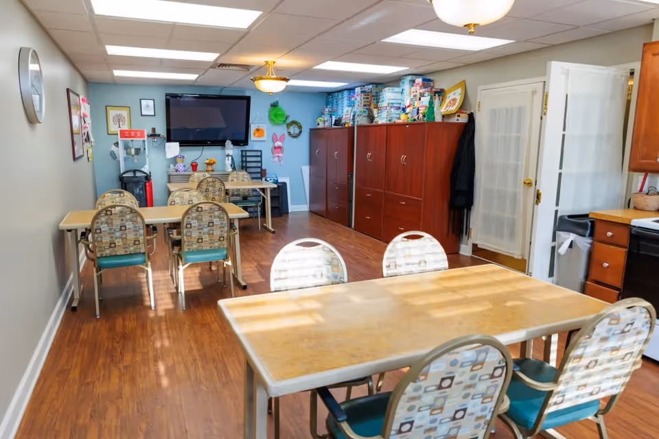 A well-lit room with wooden flooring featuring two tables with patterned chairs around them. The room has a wall-mounted TV, a popcorn machine, and several wooden cabinets filled with board games and decorations. There is a clock on the wall and a door with frosted glass panels leading to another area.