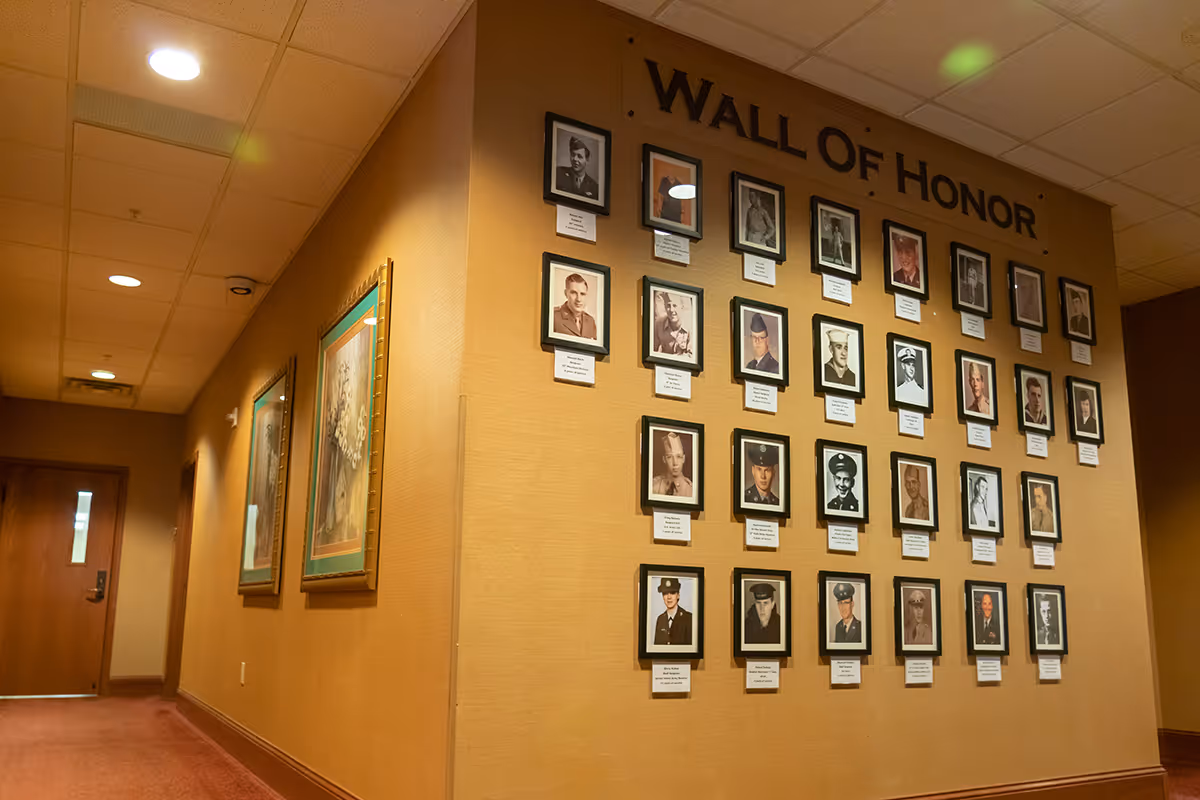 Interior hallway of a senior living facility with a 'Wall of Honor' displaying framed photographs of veterans on the right wall and two framed paintings on the left wall. The hallway has a carpeted floor and a door at the end.