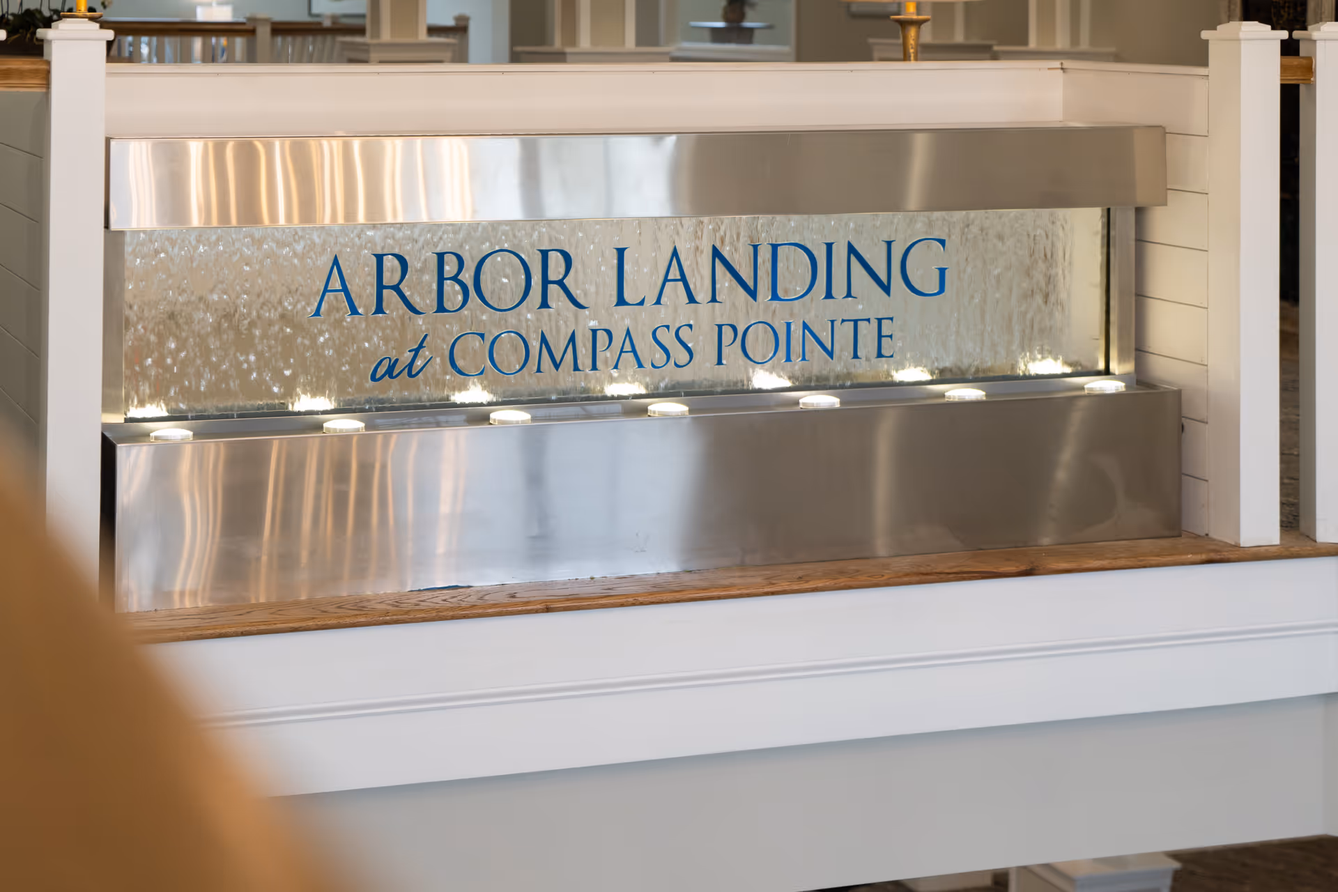 An indoor stainless-steel water feature with a glass panel sign that reads "Arbor Landing at Compass Pointe".