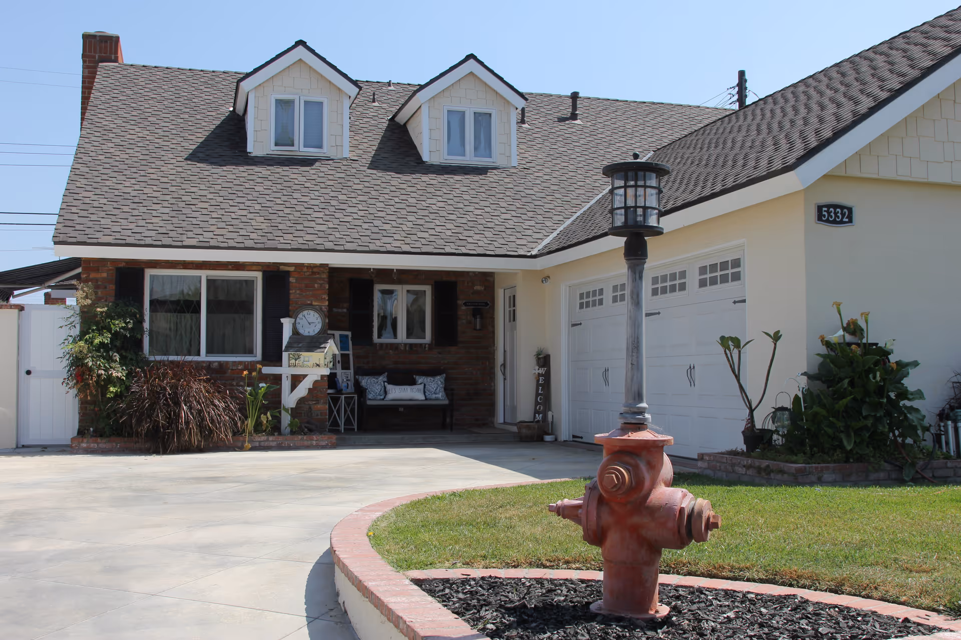 Front exterior view of a single-story house with a gray shingled roof, two dormer windows, a brick facade around the entrance, a white garage door, a red fire hydrant with a lamp post on top in the foreground, and a well-maintained lawn and driveway.