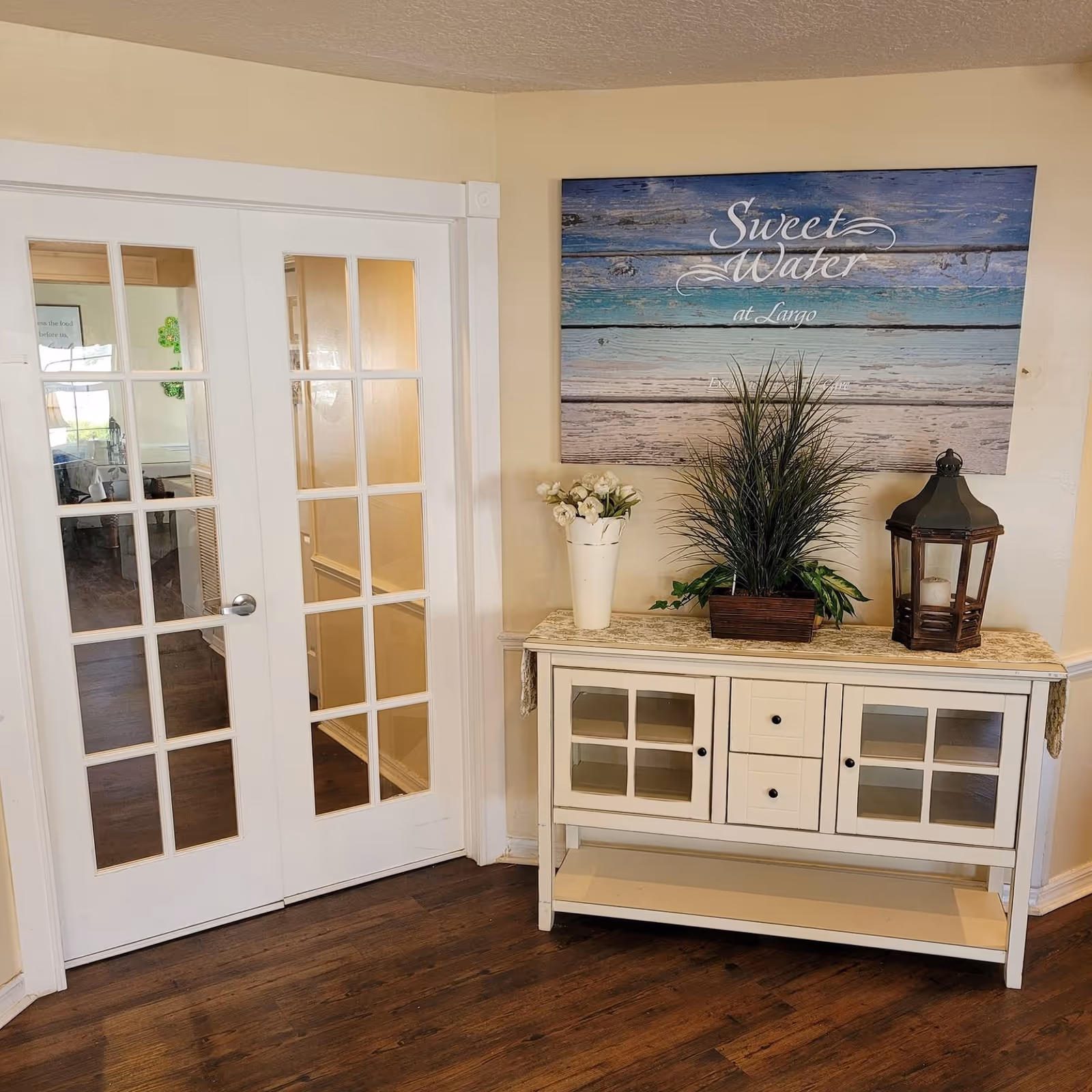 Interior corner of a room with wooden flooring, a white cabinet with glass doors and drawers, decorated with a white vase of flowers, a green potted plant, and a lantern. Above the cabinet is a wall art sign that reads 'Sweet Water at Largo'. To the left are white double doors with glass panes.