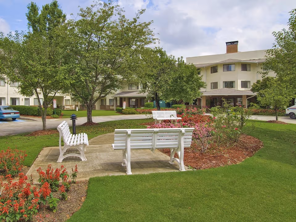 Outdoor garden area at Regency House by Barclay House featuring green grass, flower beds with red and pink flowers, several trees, white benches on a concrete path, and a multi-story building in the background under a partly cloudy sky.