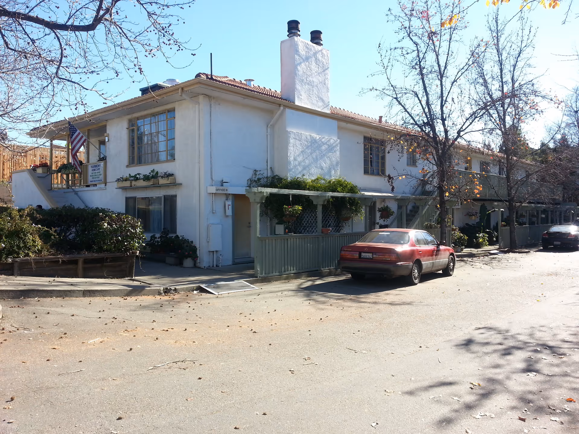 Exterior view of a two-story white building with a red-tiled roof, identified as Harmony Home Care in Walnut Creek. The building has multiple windows, a chimney, and a green wooden railing with hanging plants. Two cars are parked in front of the building on a paved area with some trees around.