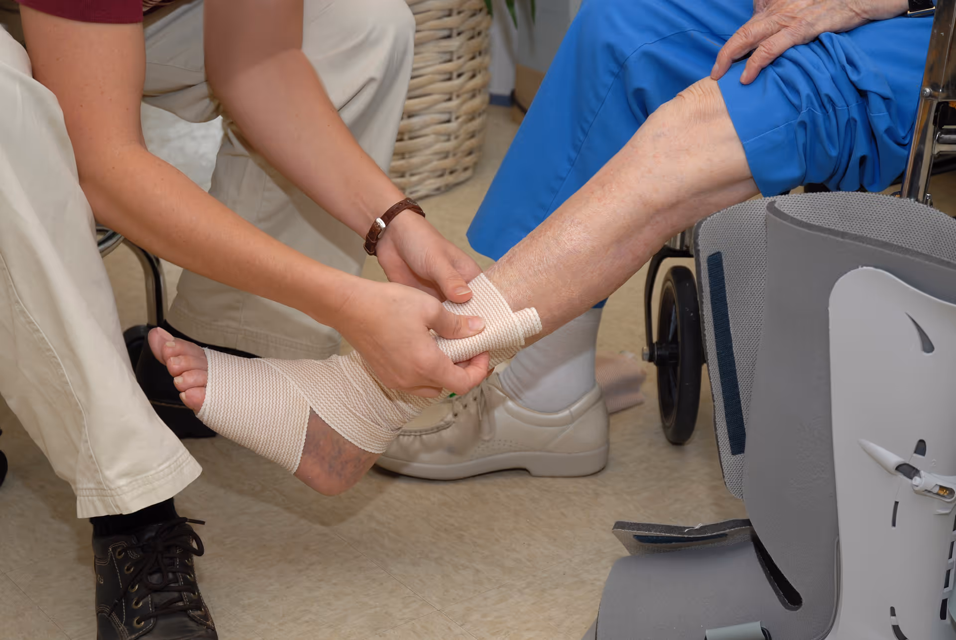 A caregiver is wrapping a bandage around the ankle of an elderly person who is seated in a wheelchair. The elderly person is wearing blue pants and beige shoes, and the caregiver is kneeling on the floor while attending to the bandage.
