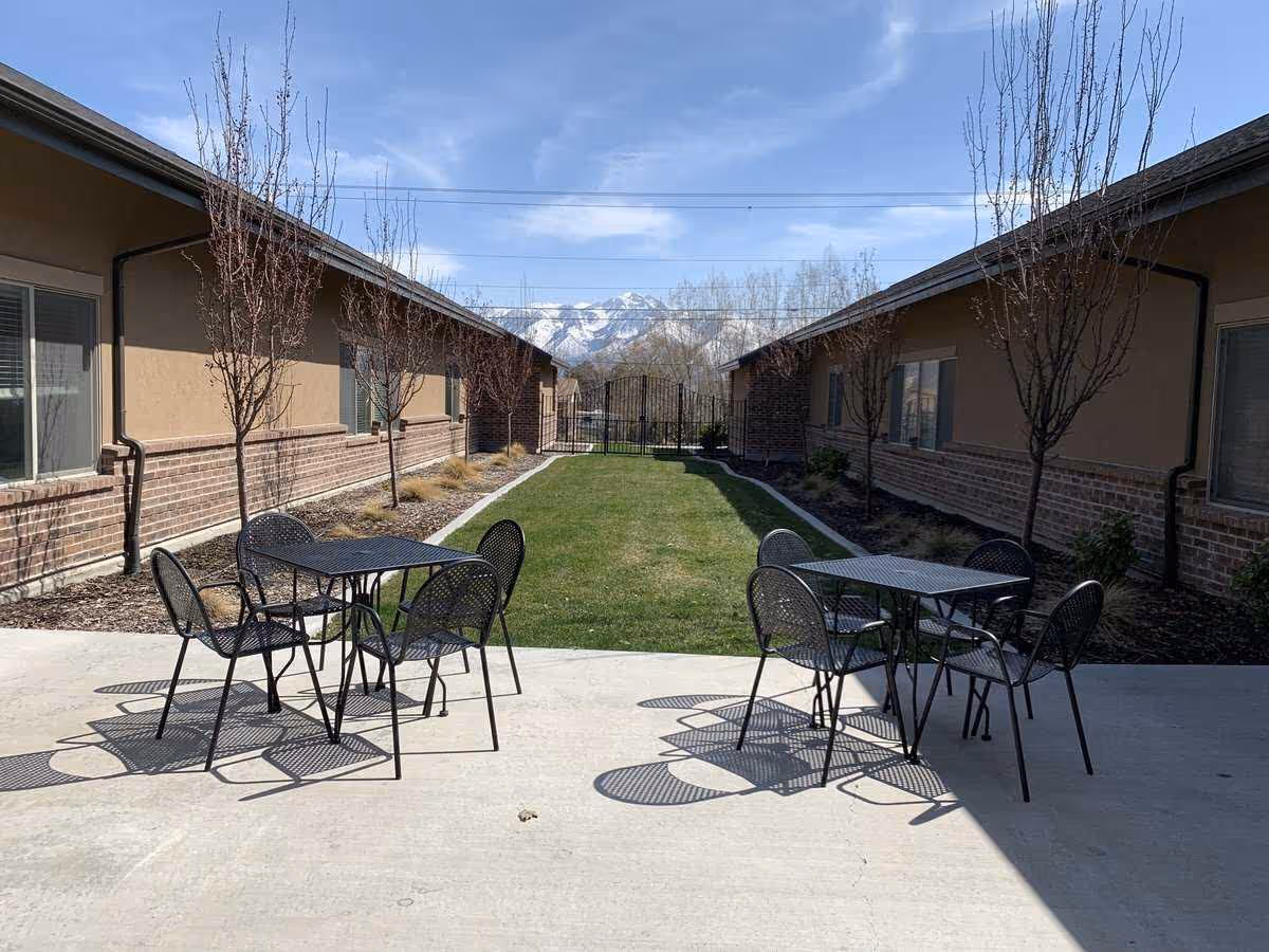 Outdoor patio area with two black metal tables and eight matching chairs on a concrete surface. The patio is flanked by two single-story buildings with beige walls and brick accents. There are small leafless trees planted along the sides of the buildings and a green grassy area in the center leading to a black metal gate. Snow-capped mountains and a partly cloudy blue sky are visible in the background.