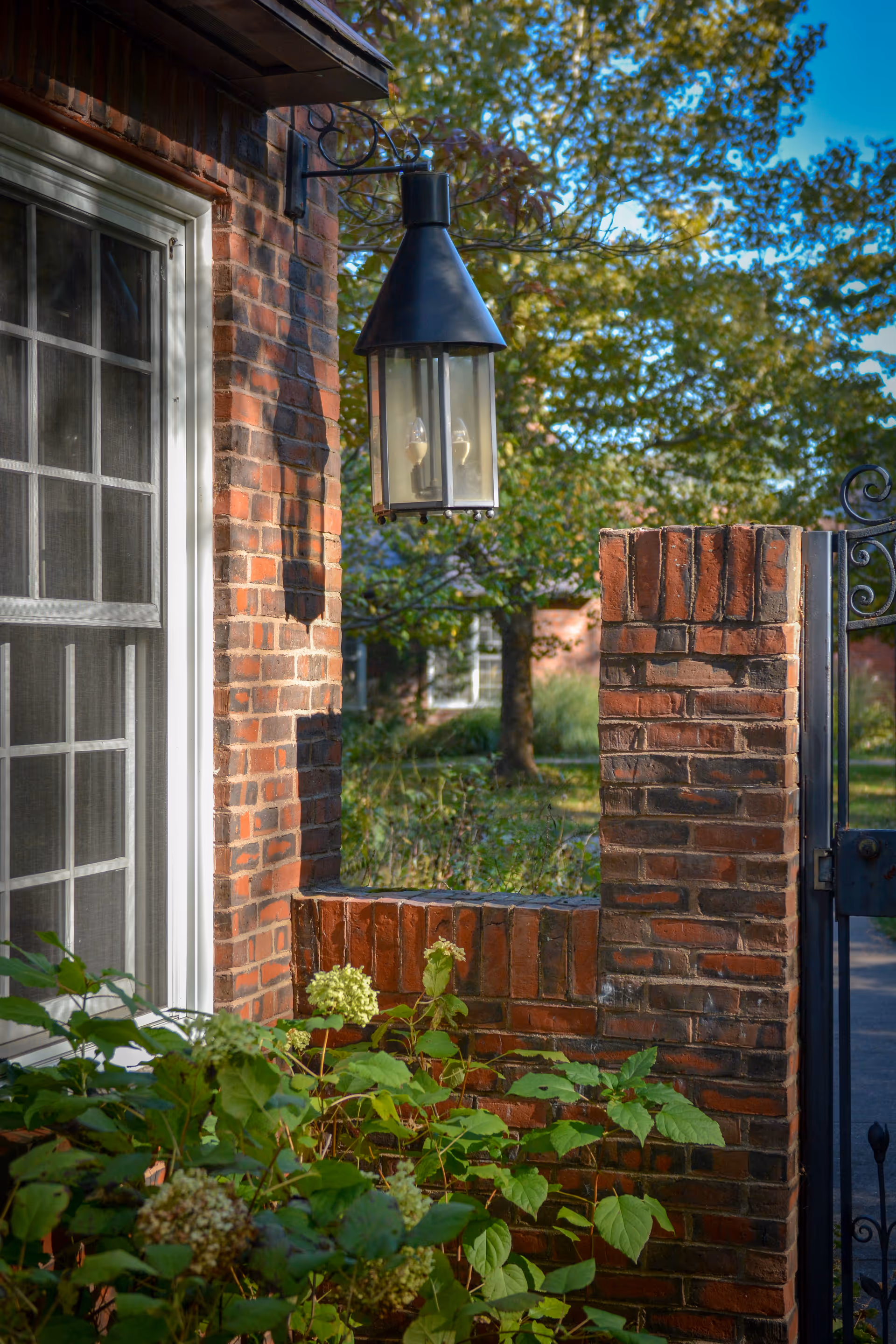 A close-up view of a brick wall and window with white trim, featuring a black outdoor lantern hanging on the wall. Green plants with some flowers are in the foreground, and trees and greenery are visible in the background under a clear blue sky.