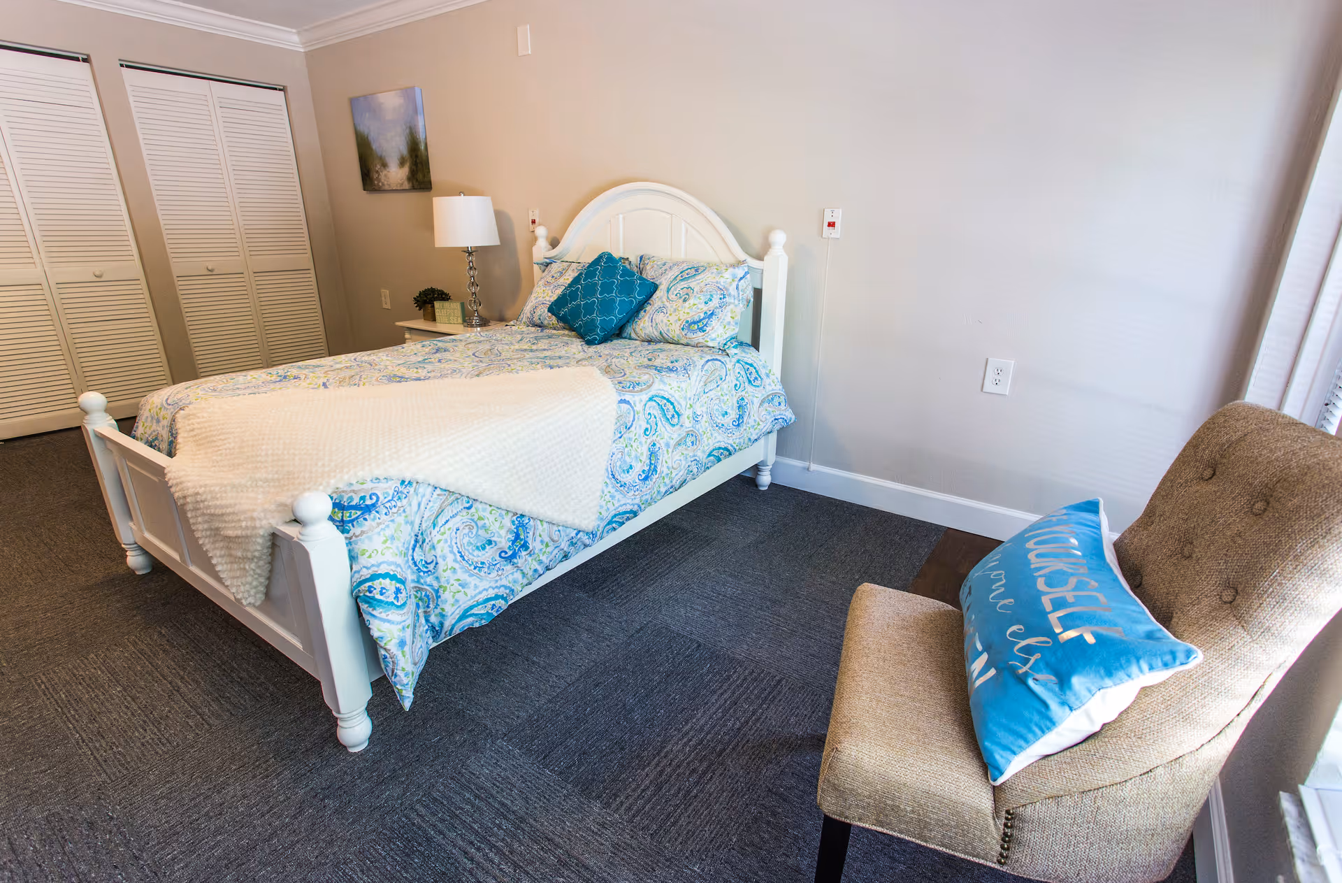 A bedroom with a white wooden bed frame featuring a blue and white paisley patterned bedspread and pillows. There is a beige upholstered chair with a blue decorative pillow next to a window. A nightstand with a lamp and a small plant is beside the bed, and a closet with white louvered doors is visible in the background.