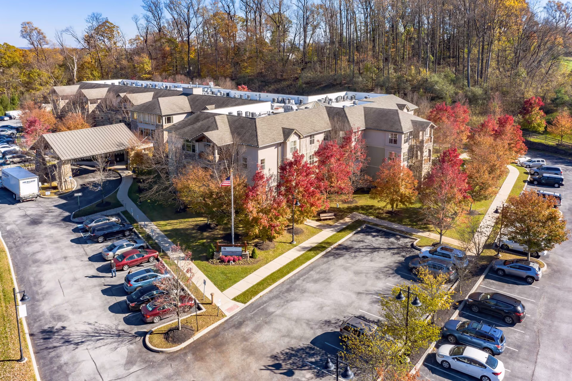Aerial view of the Spring Mill senior living facility surrounded by trees with autumn foliage. The building has a large covered entrance and is bordered by parking lots with numerous cars. Sidewalks and landscaped areas with benches and an American flag are visible in front of the building.