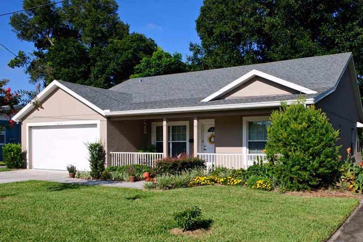 Single-story house with a gray roof, beige exterior walls, a white garage door, and a small front porch with white railing. The front yard is well-maintained with green grass, various shrubs, and flowering plants. Large trees are visible in the background under a clear blue sky.