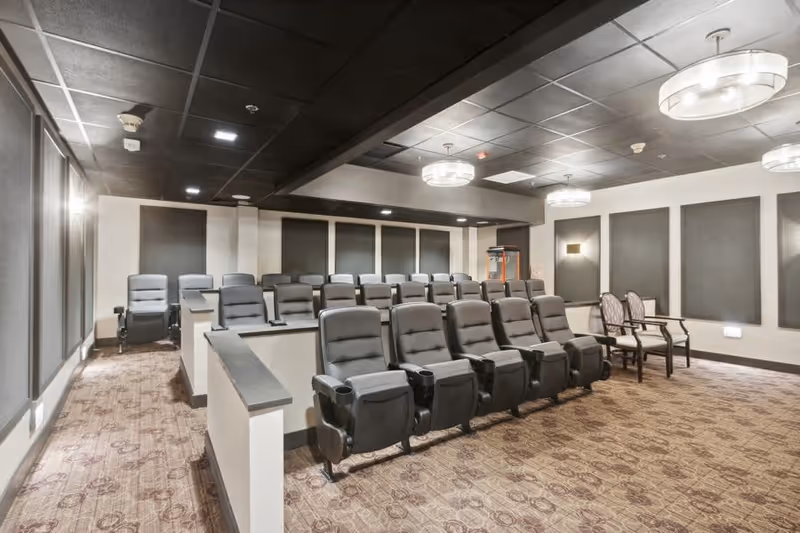A small theater room with multiple rows of black reclining chairs arranged in tiers, patterned carpet flooring, and soft lighting from ceiling fixtures. The walls have dark acoustic panels, and there is a popcorn machine in the back corner.