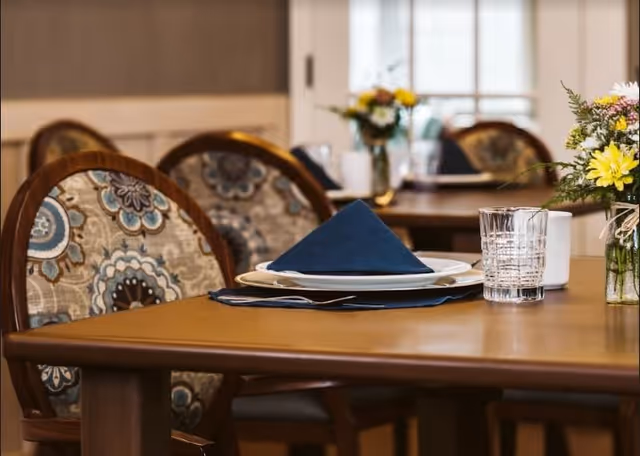 A close-up view of a dining table set with a white plate, a folded dark blue napkin, a glass of water, and a small vase with yellow and white flowers. The table is surrounded by chairs with patterned upholstery featuring blue, beige, and brown colors. In the background, more tables and chairs are visible with similar settings and floral arrangements.