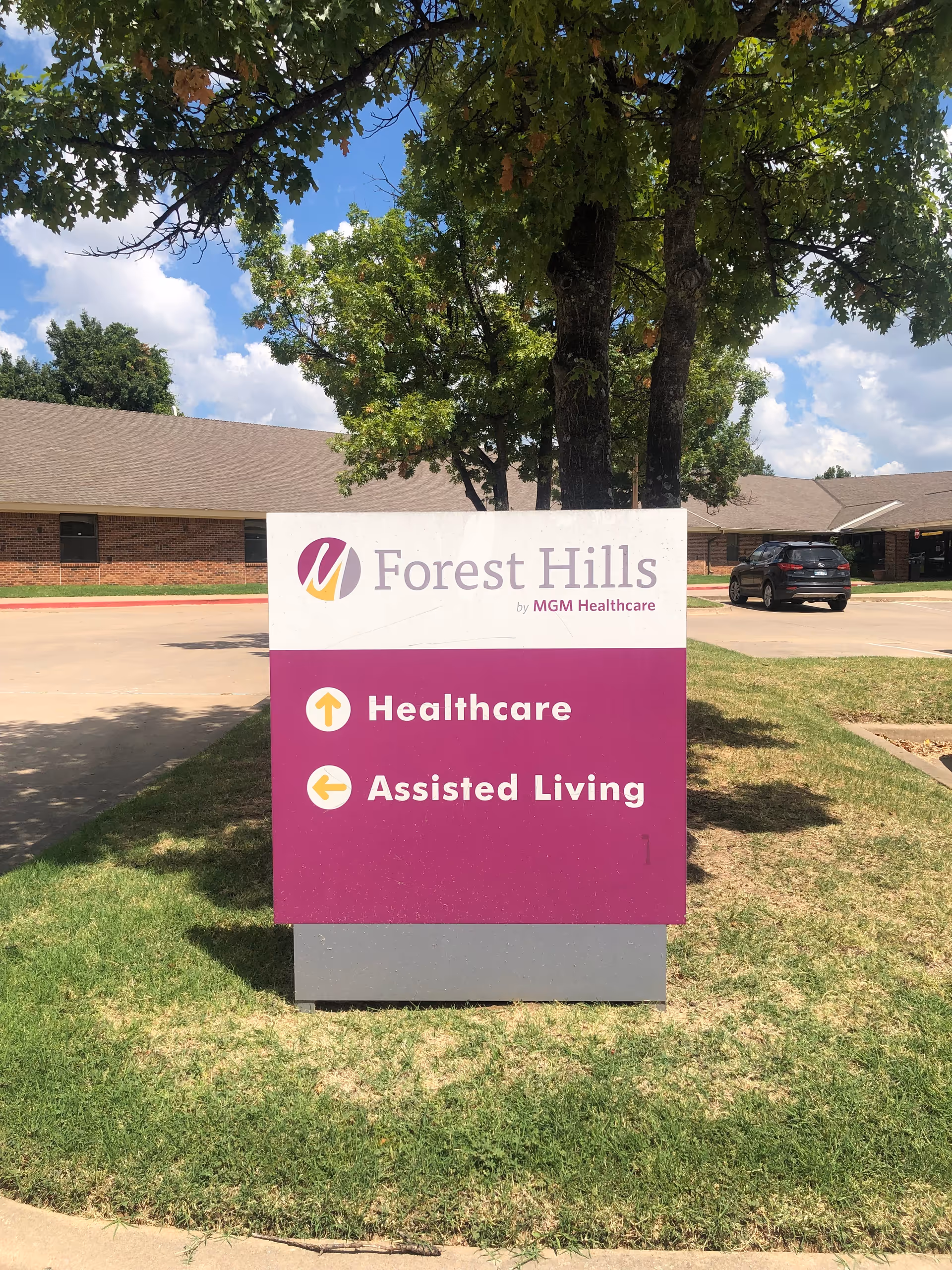 Outdoor image of a sign for Forest Hills by MGM Healthcare, indicating directions for Healthcare (straight ahead) and Assisted Living (to the left). The sign is placed on a grassy area with a tree behind it, and a building and parked car are visible in the background under a partly cloudy sky.