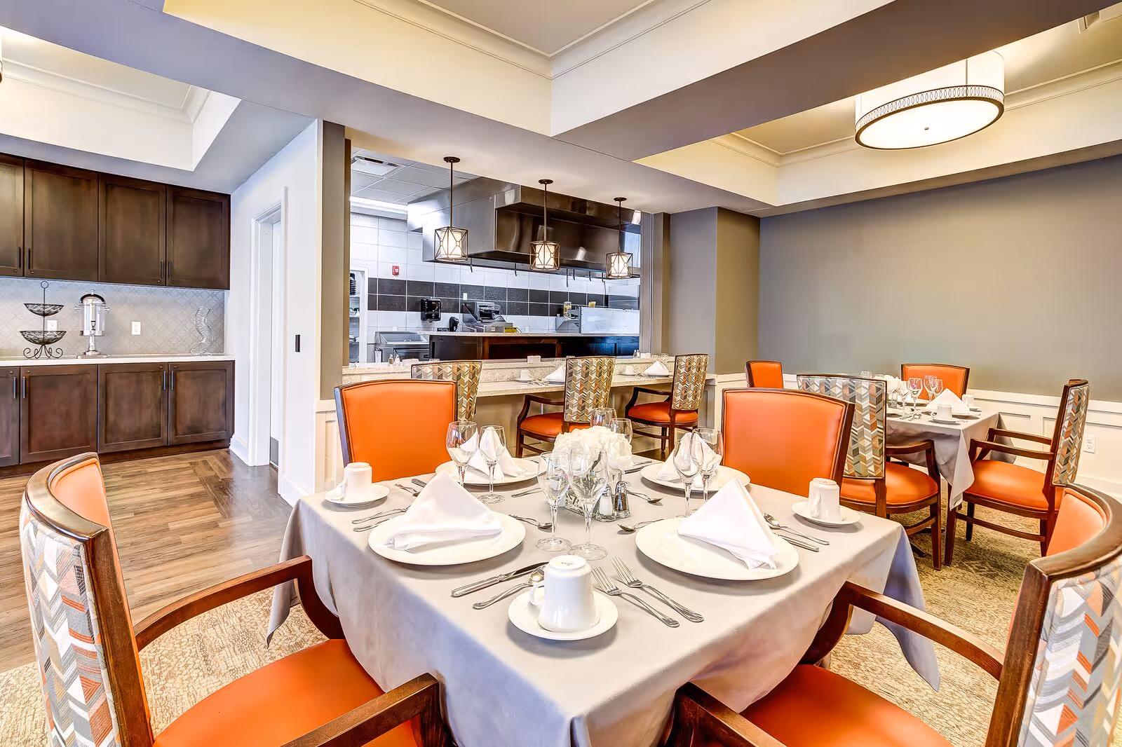 A dining room with tables set for a meal, featuring white plates, folded napkins, glassware, and silverware. The chairs have orange cushions and patterned backs. In the background, there is a view into a kitchen area with stainless steel appliances and three pendant lights hanging above a counter.