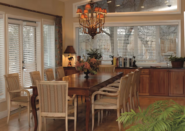 A dining room with a long wooden table surrounded by upholstered chairs. The room has large windows with white blinds, a chandelier with multiple lampshades hanging above the table, and a sideboard with plants and books. There is also a table lamp on a small cabinet in the corner.