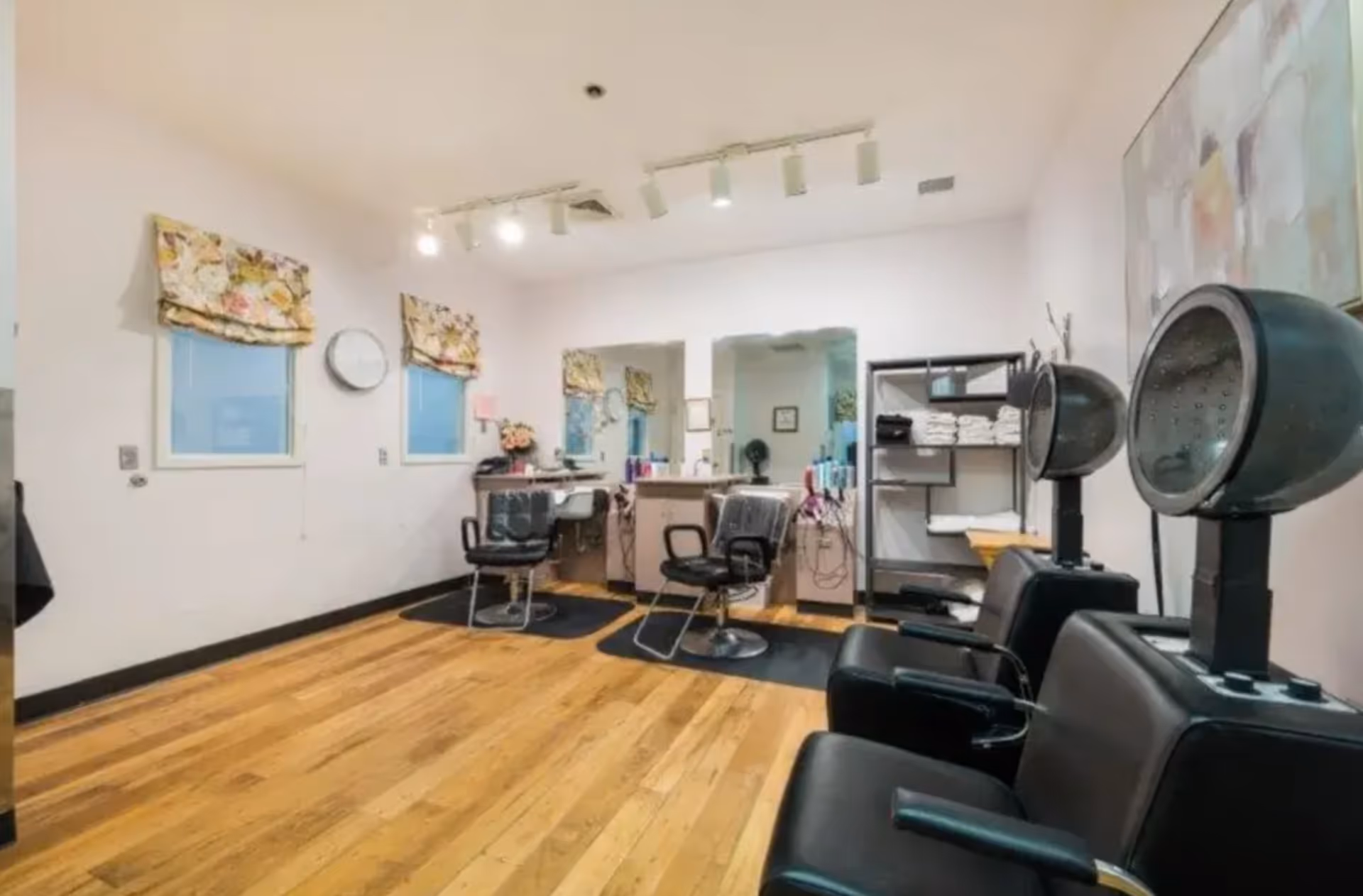 Interior view of a hair salon area with wooden floors, two black salon chairs in front of mirrors and counters with hair products, and two black hair dryer chairs on the right side. There are floral curtains on the windows and shelves with towels and supplies.