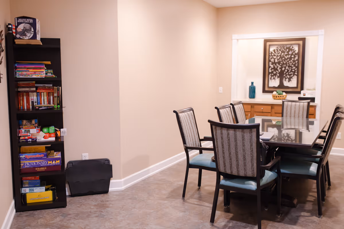 A well-lit room with a rectangular glass-top table surrounded by six upholstered chairs with patterned backs and blue seats. To the left, there is a tall black bookshelf filled with board games, books, and other items. On the far wall, there is a built-in counter with wooden cabinets underneath, decorated with a blue vase, a basket, and a framed artwork of a tree.