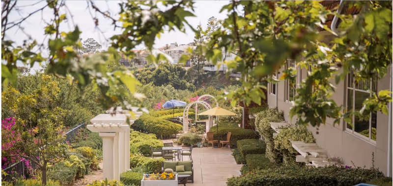 A landscaped outdoor patio area at Crown Cove featuring seating arrangements with chairs and tables, umbrellas for shade, lush green bushes, trees, and flowering plants. The patio is adjacent to a building with large windows, and there are decorative white pergolas and arches along the walkway.