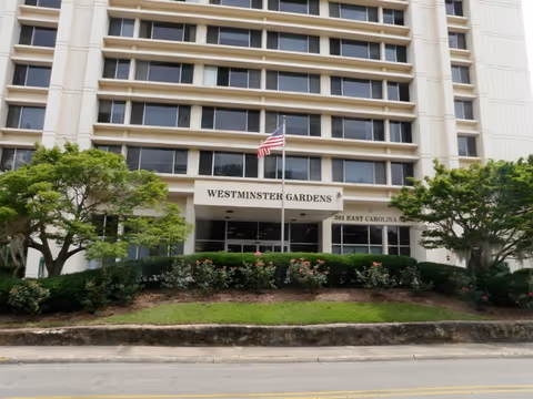 Front exterior view of a multi-story building named Westminster Gardens with an American flag on a flagpole in front, surrounded by green bushes and trees.