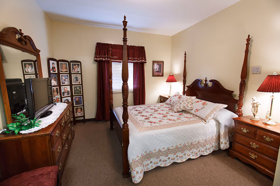 A cozy bedroom with a wooden four-poster bed covered in a white quilt with floral patterns. There are two matching wooden nightstands with red lamps on either side of the bed. A wooden dresser with a large mirror and a TV on top is on the left side of the room. A photo collage frame with multiple pictures is placed near the window, which has red curtains.