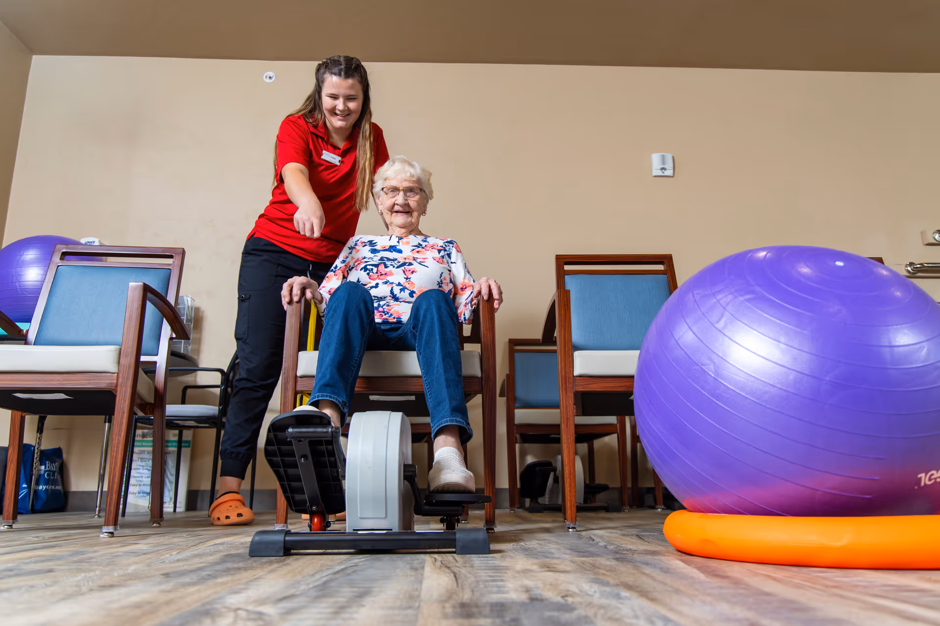 An elderly woman sitting on a chair using a pedal exerciser while a caregiver in a red shirt stands beside her, smiling and pointing. The room has wooden chairs with blue cushions and a large purple exercise ball on an orange base on the floor.