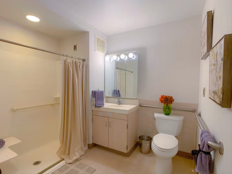 A clean and well-lit bathroom featuring a walk-in shower with a beige curtain, a vanity with a sink and mirror above it, a toilet with a green vase holding orange flowers on top, purple towels hanging on grab bars, and two framed floral artworks on the wall.