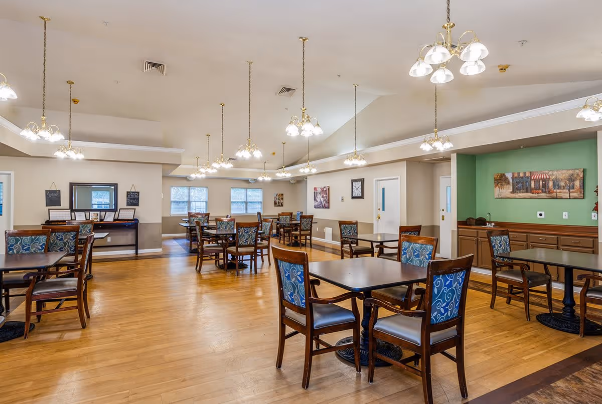 A spacious dining room with multiple wooden tables and chairs featuring blue patterned cushions. The room has hardwood floors, beige walls with a green accent wall, and several hanging light fixtures with multiple bulbs. There are windows along the far wall letting in natural light, and artwork and clocks decorate the walls.