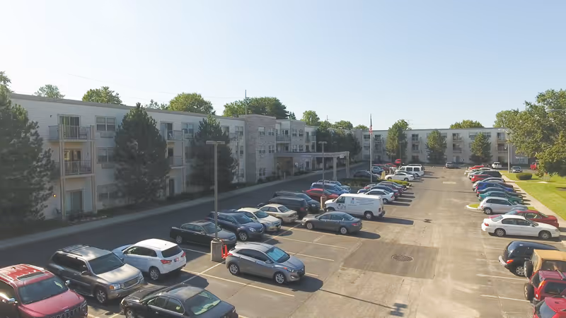 A large parking lot filled with cars in front of a three-story senior living facility building with balconies and trees around the perimeter under a clear sky.