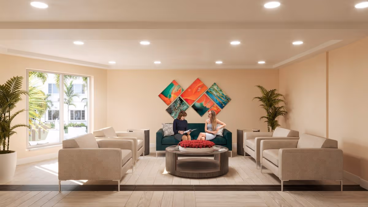 Communal lounge with sofas and armchairs arranged around a round coffee table, decorative wall art, potted plants, and two people seated on a central sofa.